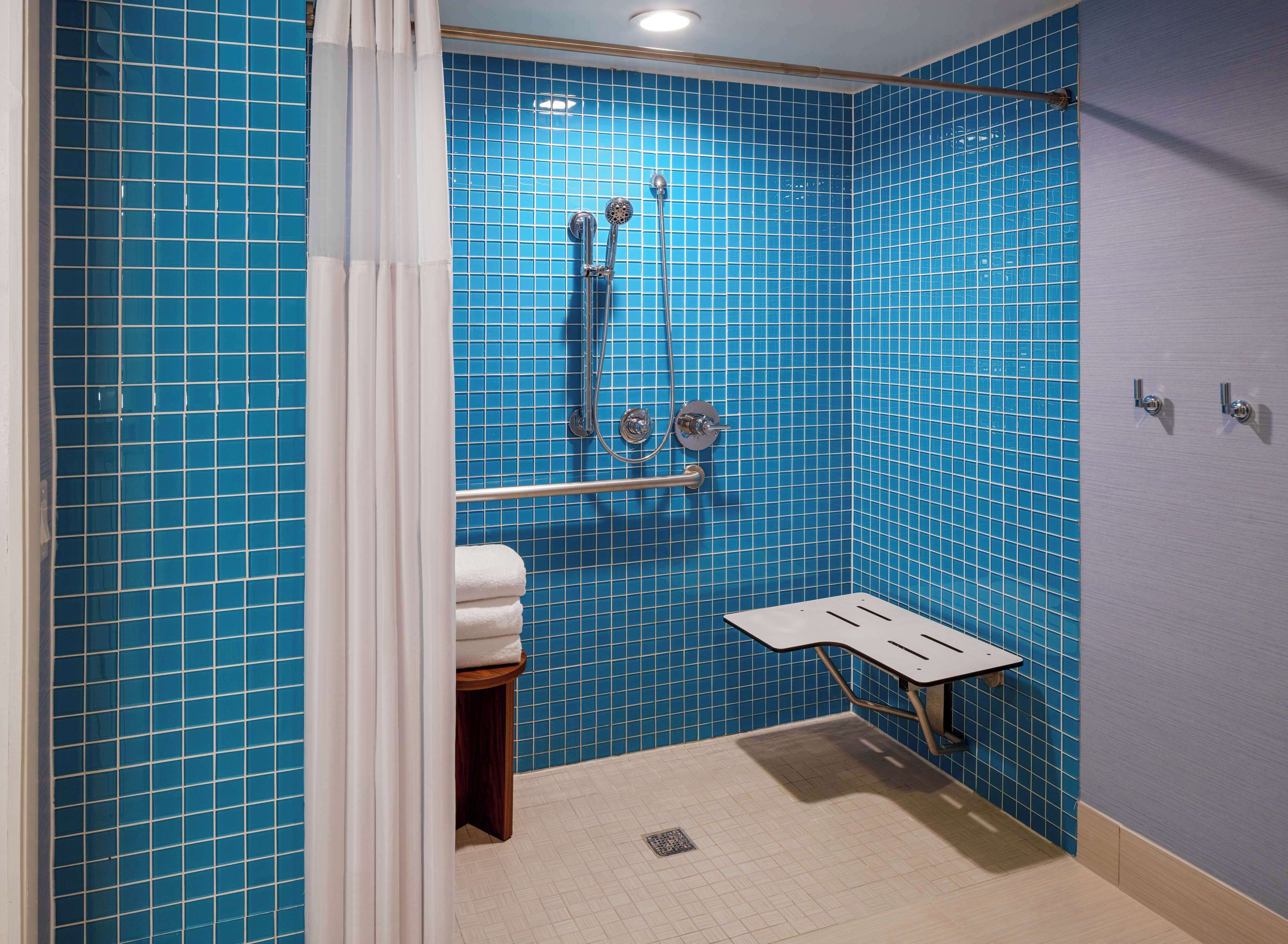 A bathroom with blue-tiled walls, a shower with a handheld showerhead, a towel rack, and a small stool.