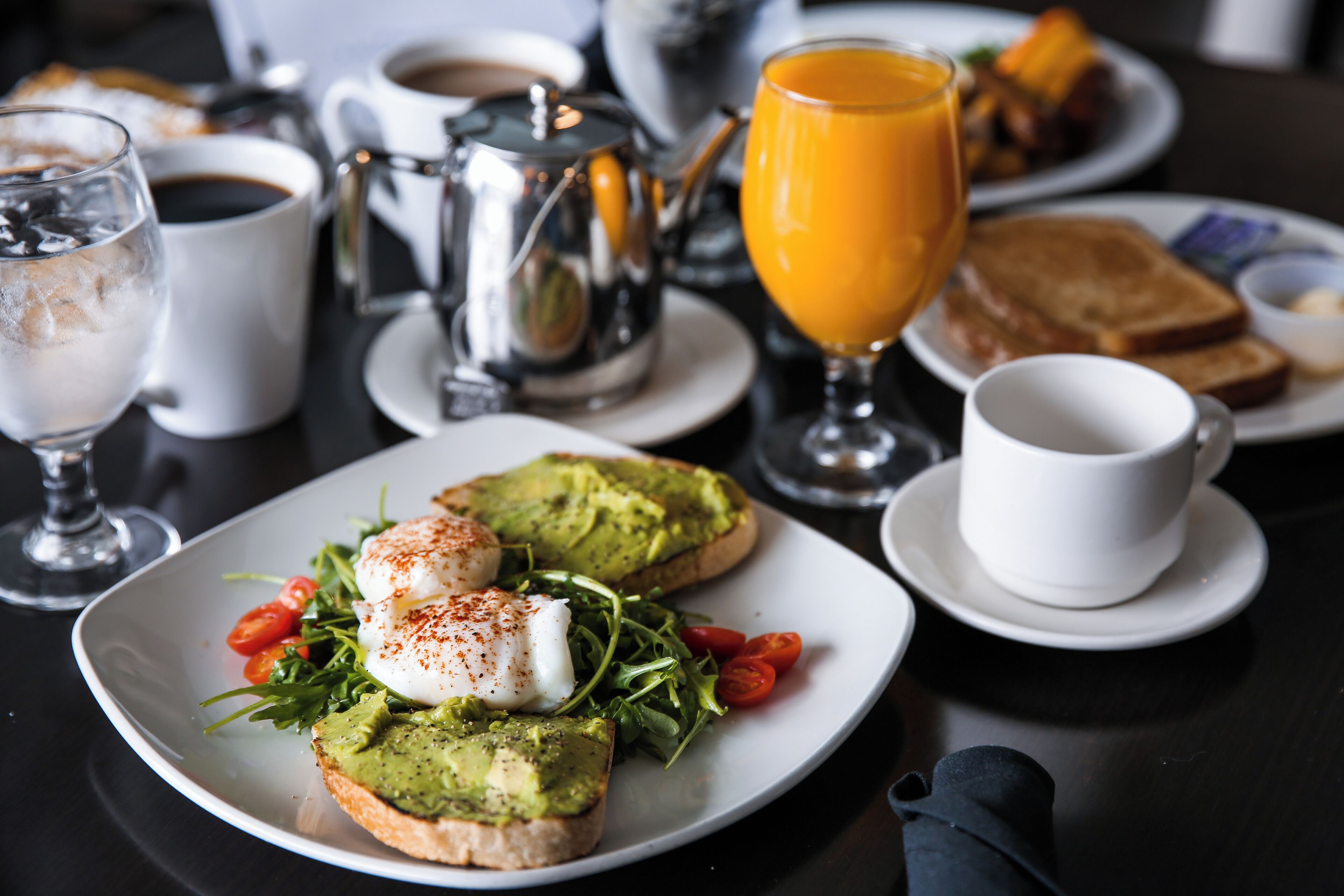 A breakfast setting with toast topped with avocado and poached eggs, a glass of orange juice, and a coffee pot.
