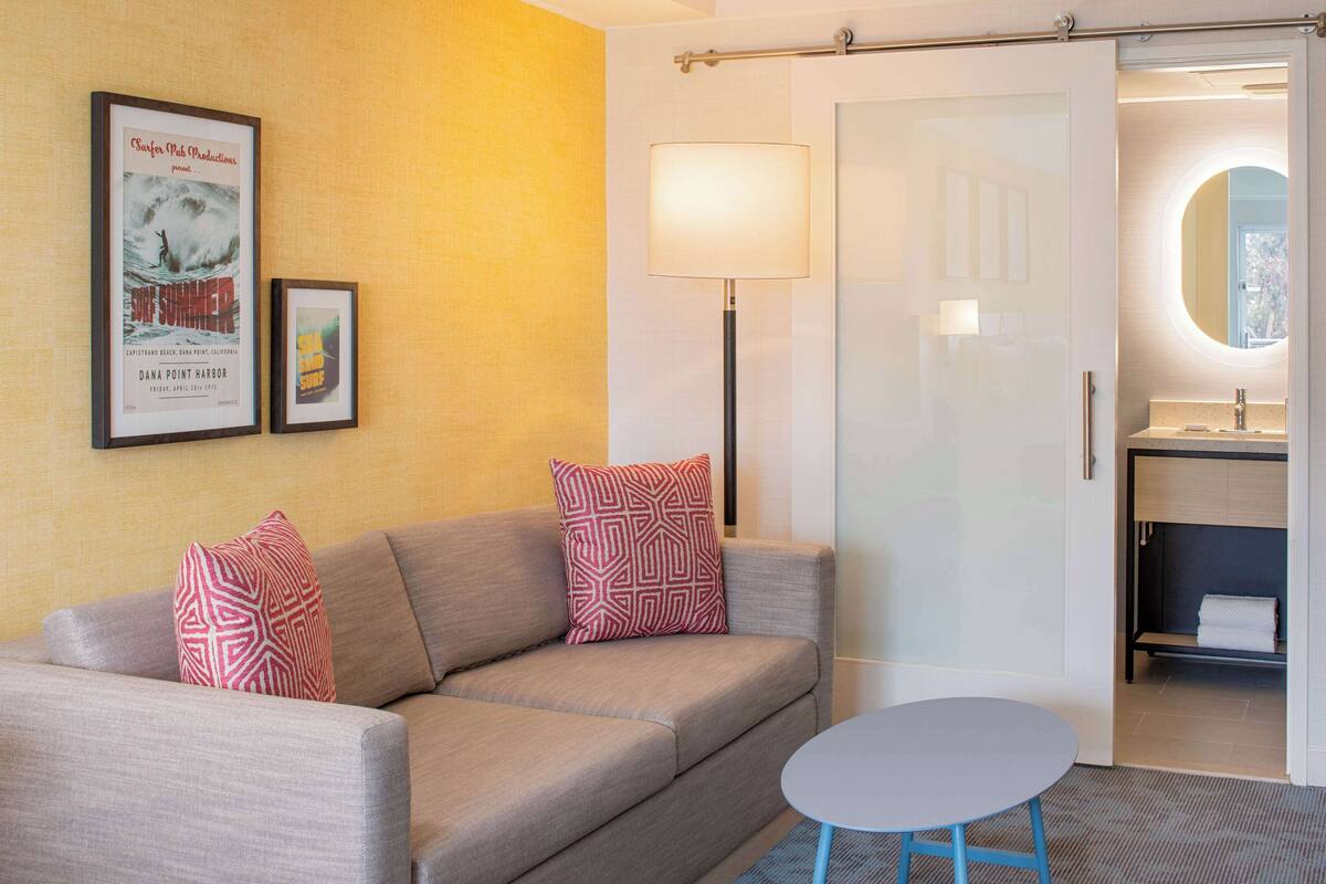 Ett hotellrum med en soffa, två mönstrade kuddar, ett litet bord, en golvlampa och en dörr som leder till ett badrum.