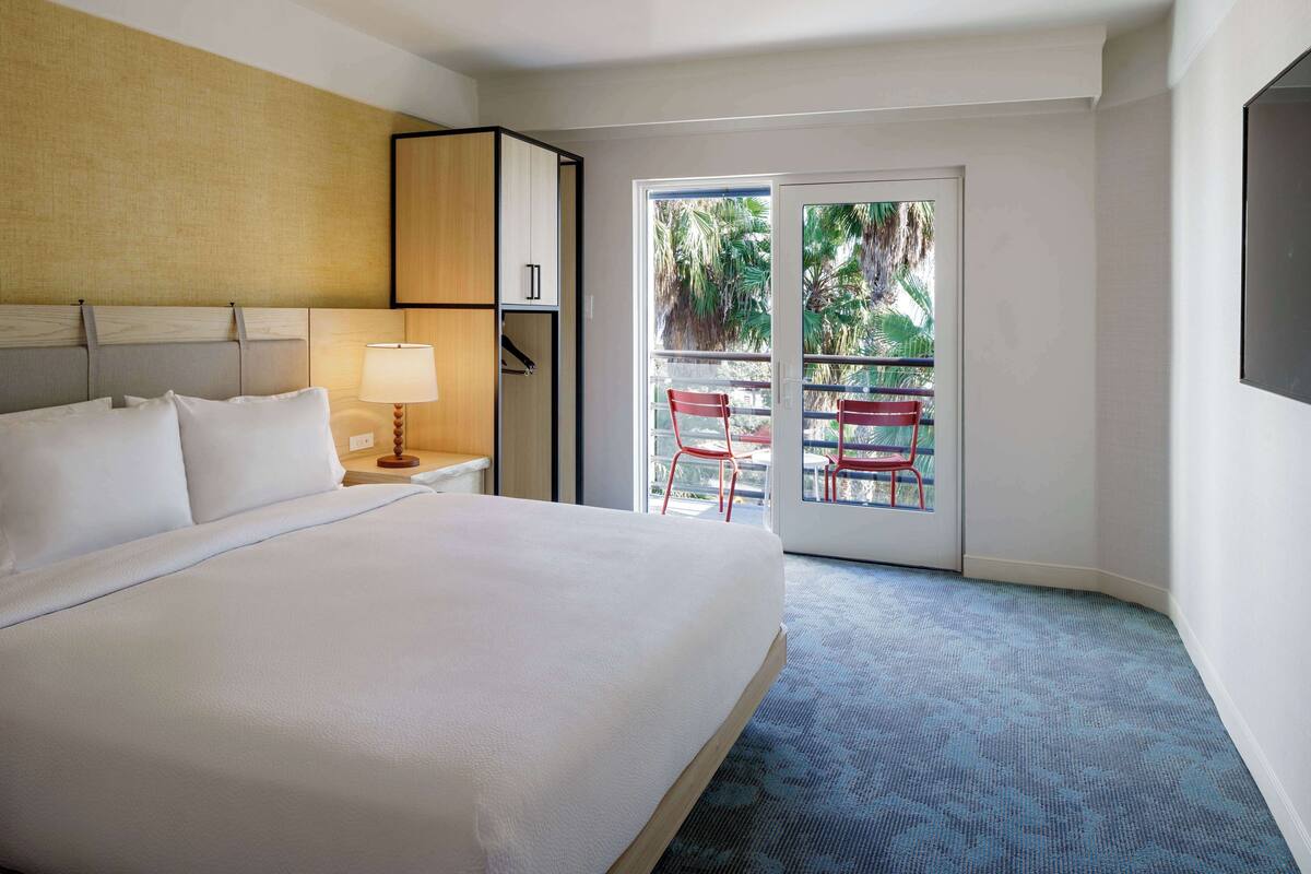 A hotel room with a large bed, a nightstand with a lamp, a wardrobe, and a balcony with red chairs.