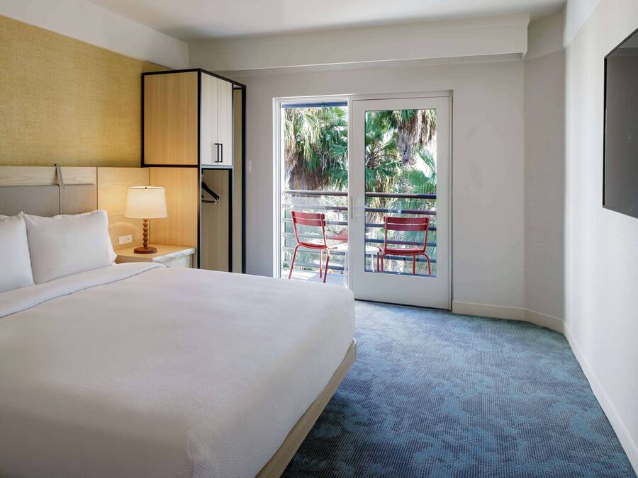 A hotel room with a large bed, a nightstand with a lamp, a wardrobe, and a balcony with red chairs.