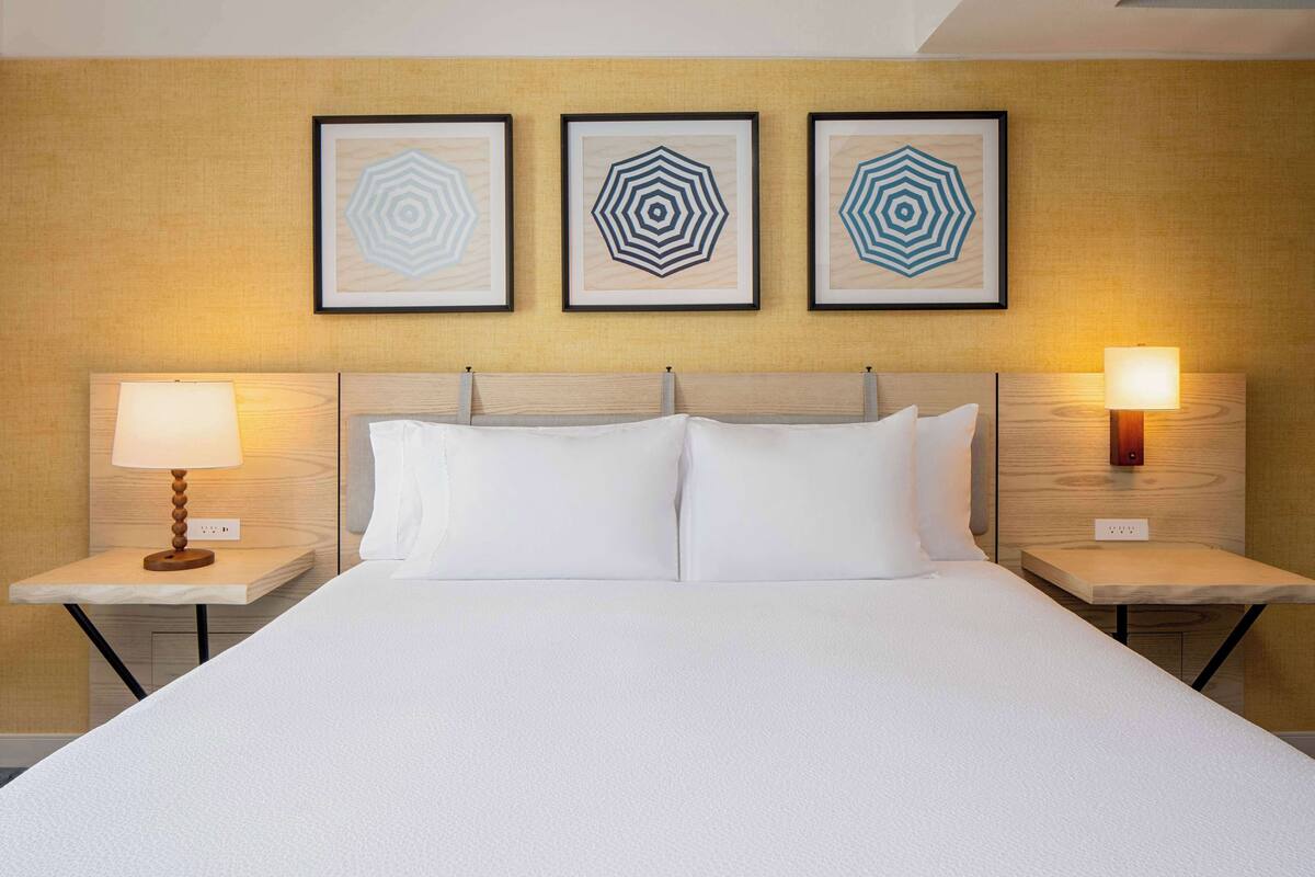 A neatly made bed with white linens, two bedside tables with lamps, and three framed geometric wall art pieces above the bed.