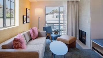 Una habitación de hotel moderna con sofá, sillón, reposapiés y una mesita. Hay una chimenea y un ventanal con cortinas.