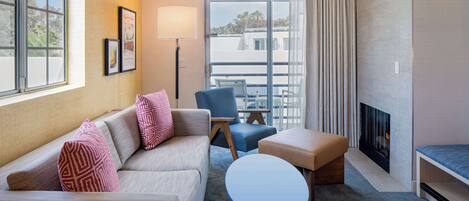 A modern hotel room with a sofa, armchair, ottoman, and a small table. There is a fireplace and a large window with curtains.