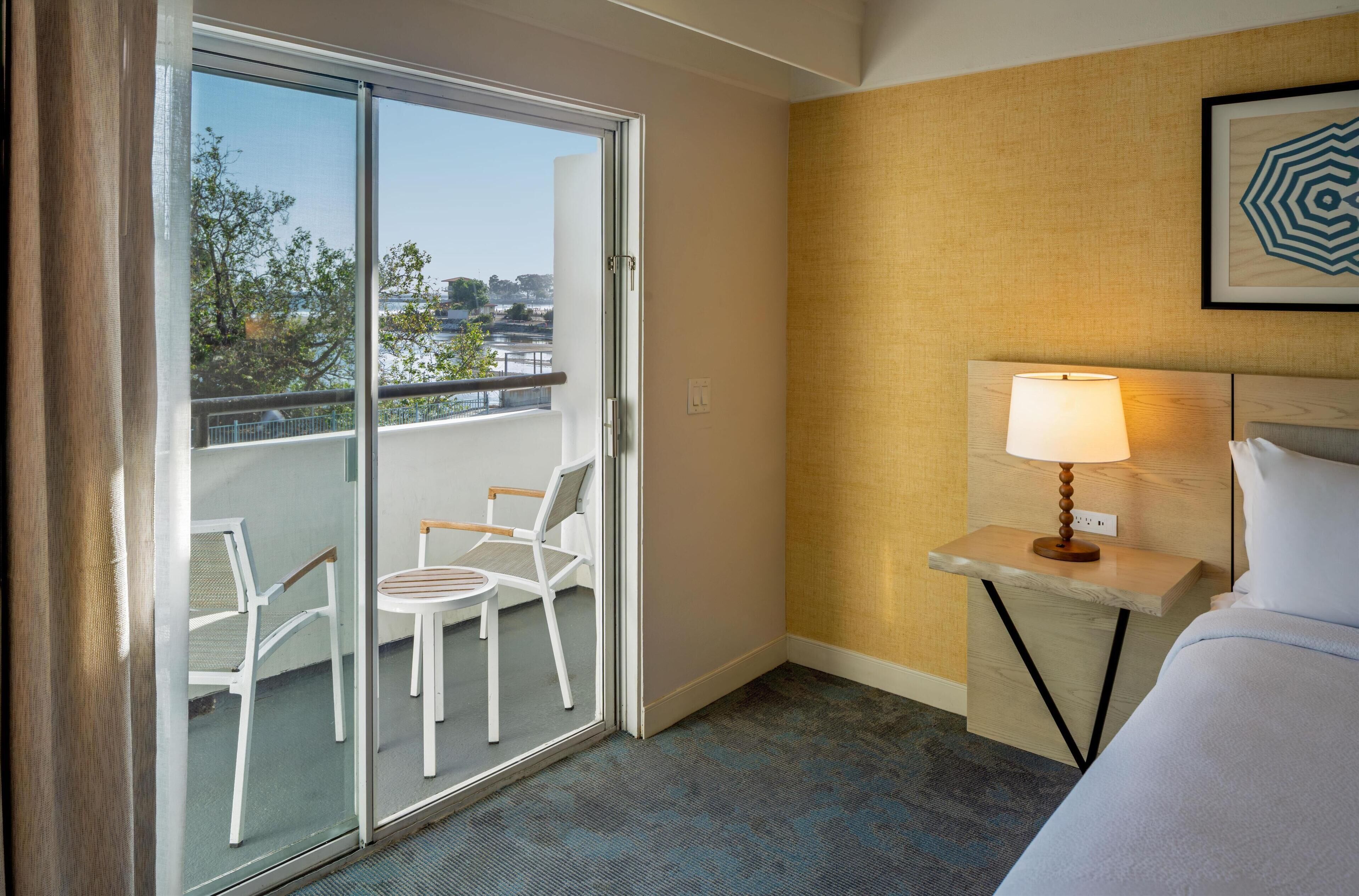 Habitación de hotel con cama, mesita de noche con lámpara, una silla y balcón con sillas.