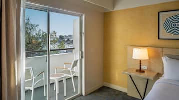 Habitación de hotel con cama, mesita de noche con lámpara, una silla y balcón con sillas.