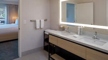 A hotel bathroom with two sinks, a large mirror, and a bed visible through an open door.