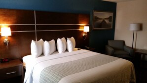 Ein Hotelzimmer mit einem großen Bett, zwei Nachttischlampen, einem Nachttisch mit einer Uhr, einem Sessel und einem gerahmten Bild an der Wand.