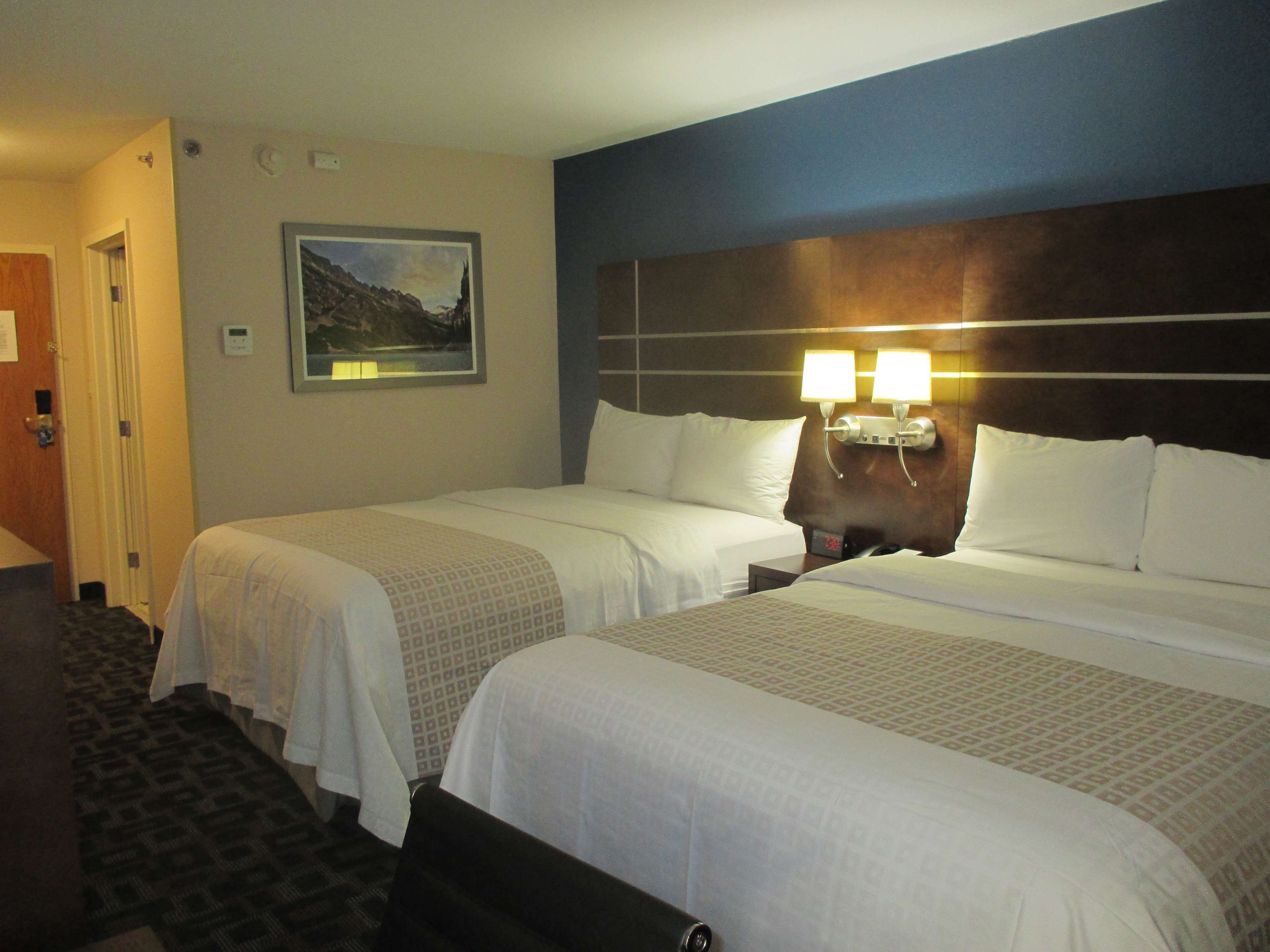 A hotel room with two beds, a nightstand with a clock, a painting on the wall, and a door to the left.