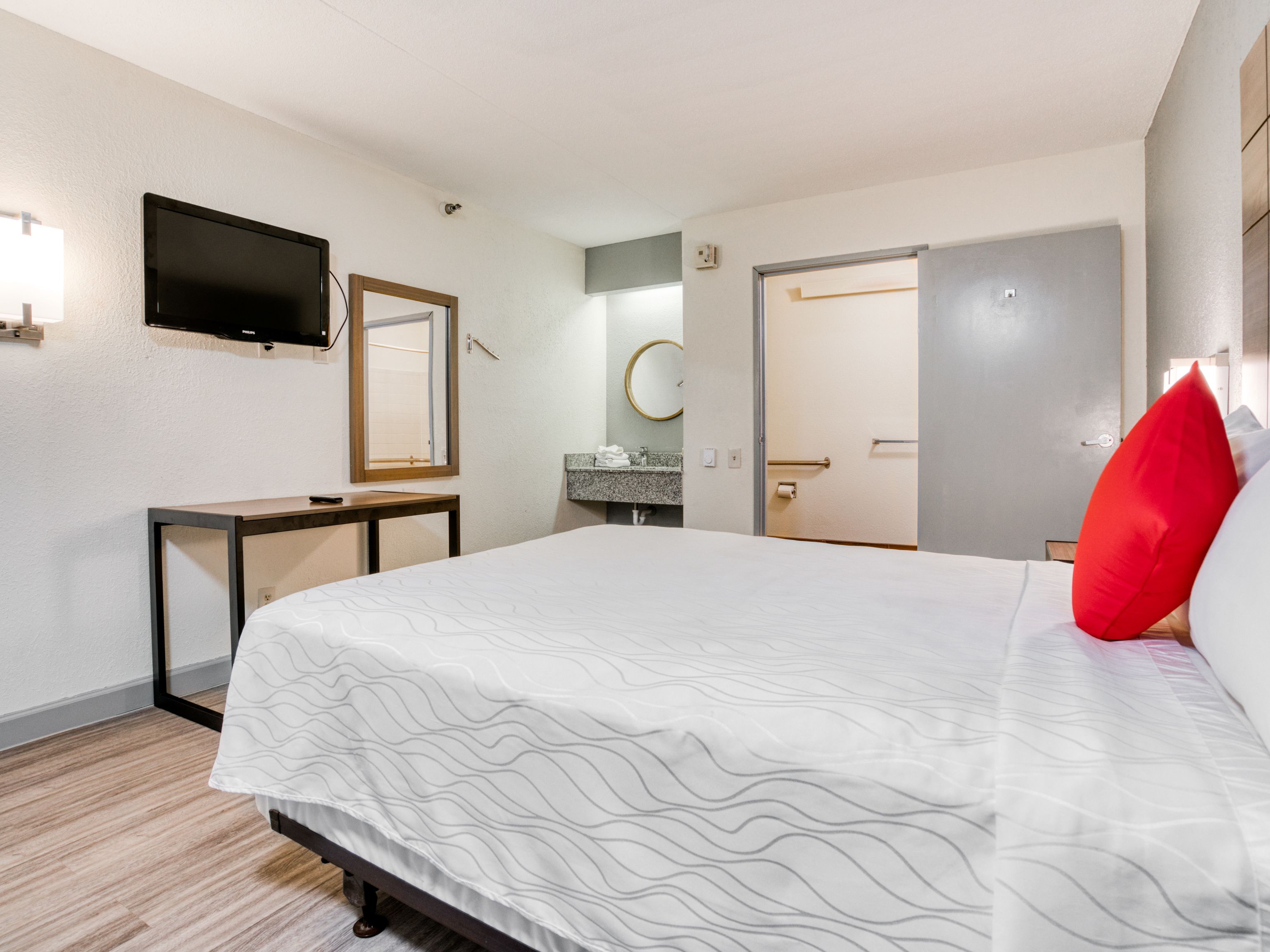 Une chambre d’hôtel avec un lit, un téléviseur fixé au mur, une table de chevet, un miroir, une porte de salle de bain et un oreiller rouge.