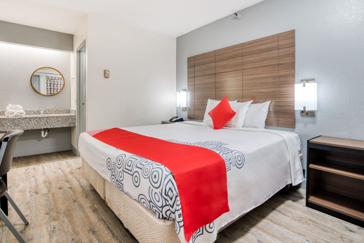 Quarto de hotel com cama, mesa de cabeceira, espelho, toucador no banheiro e piso de madeira.