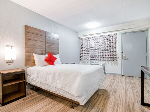A hotel room with a wooden headboard, a bedside table, a red pillow, a white bedspread, a window with patterned curtains, and a door with a lock.