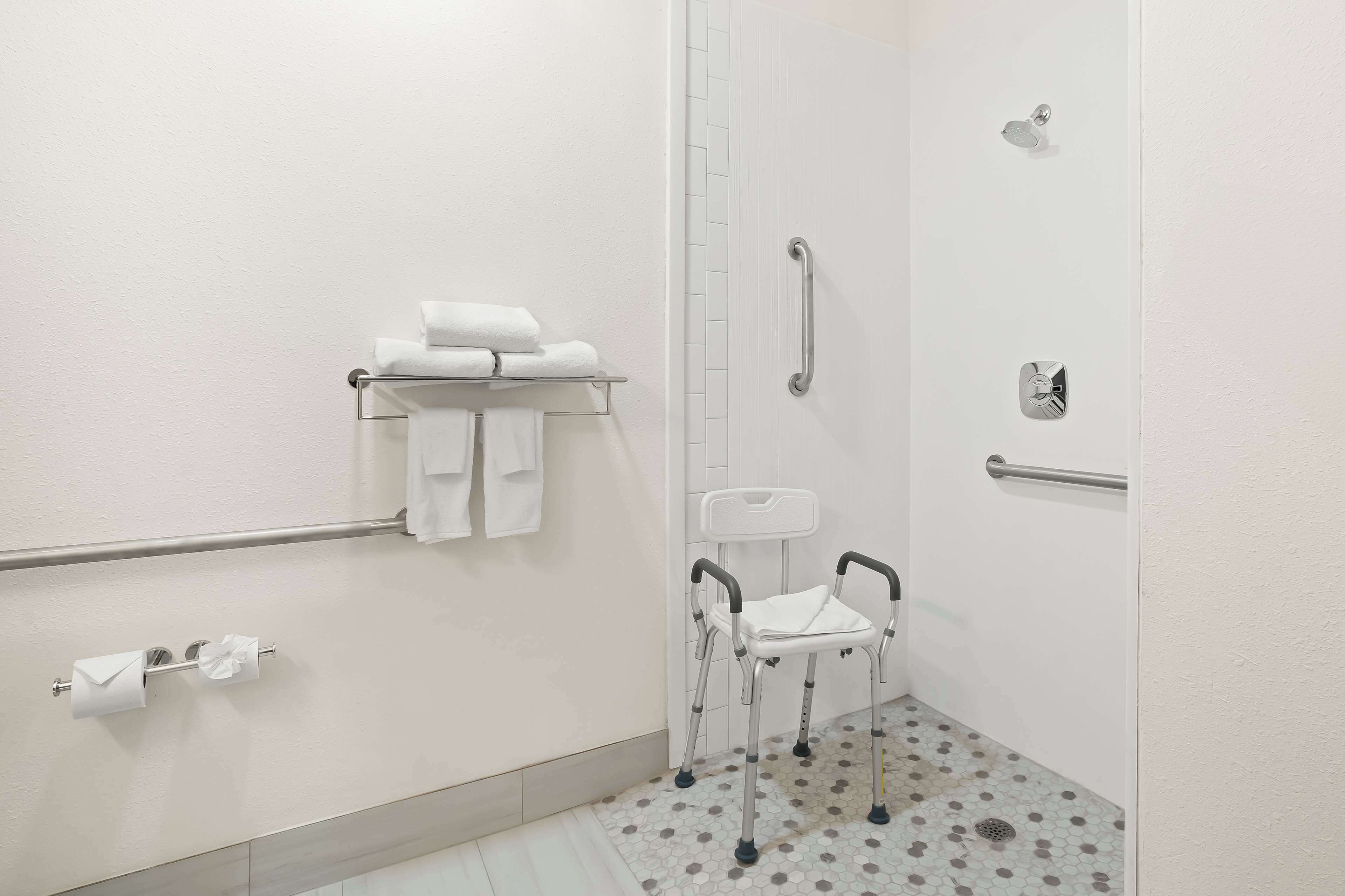 Un baño con suelo de azulejos blancos, un inodoro blanco, un lavabo blanco, un perchero blanco, una silla de ducha blanca y un asidero blanco.