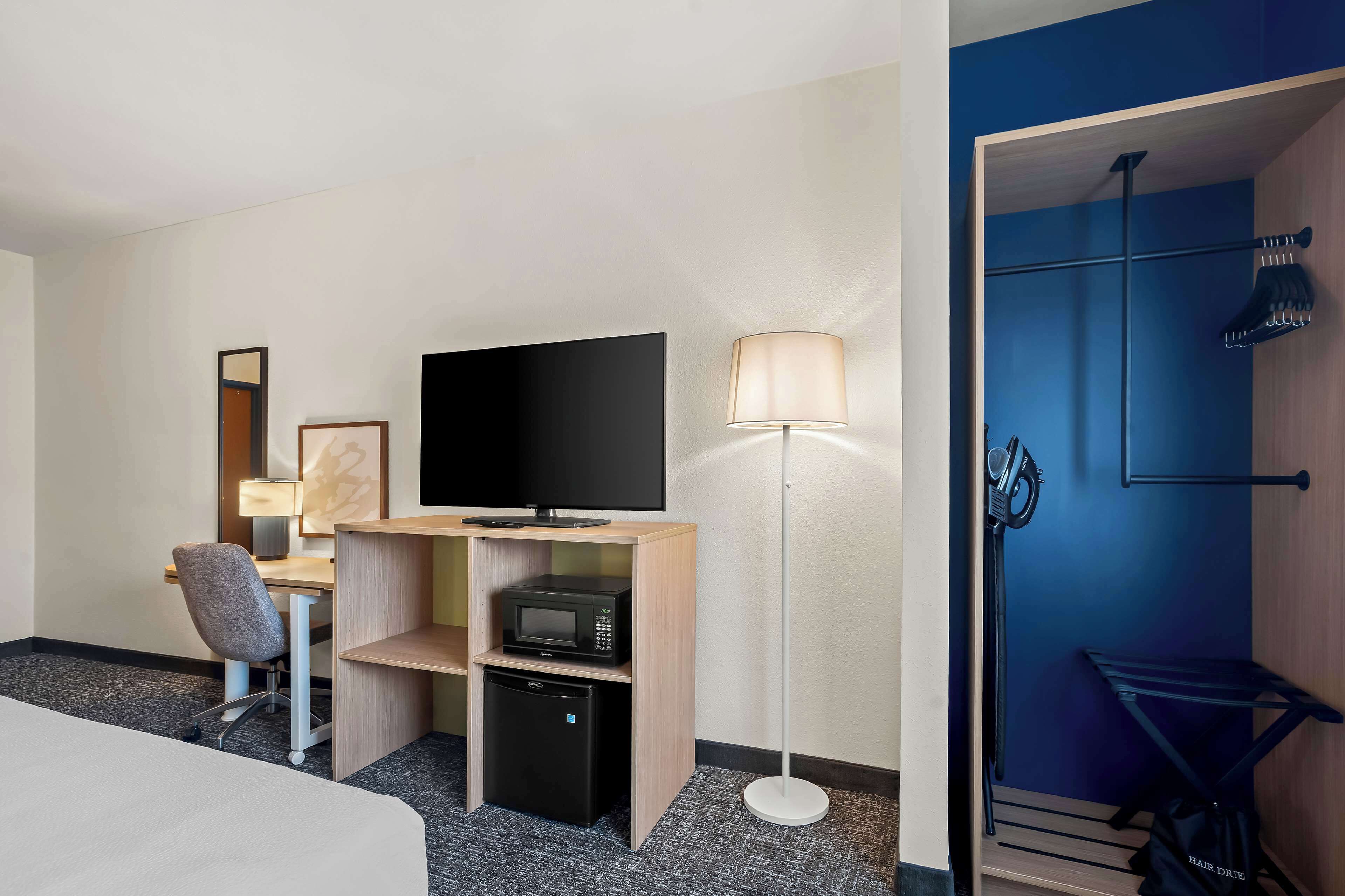 Une chambre d’hôtel comprenant un lit, un bureau avec une chaise, un téléviseur à écran plat, un four à micro-ondes, un mini-réfrigérateur, une lampe et une porte bleue avec un porte-manteau.