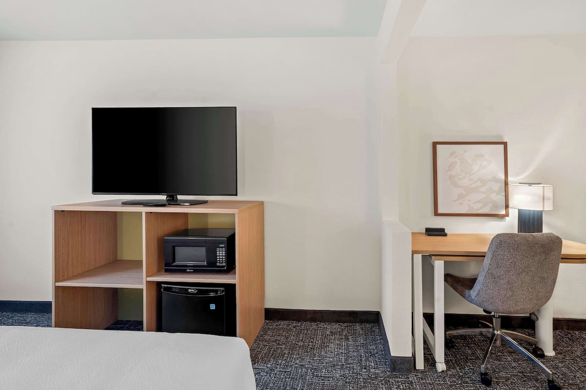 Una habitación de hotel con cama, escritorio con computadora, televisión, microondas y un mueble pequeño.