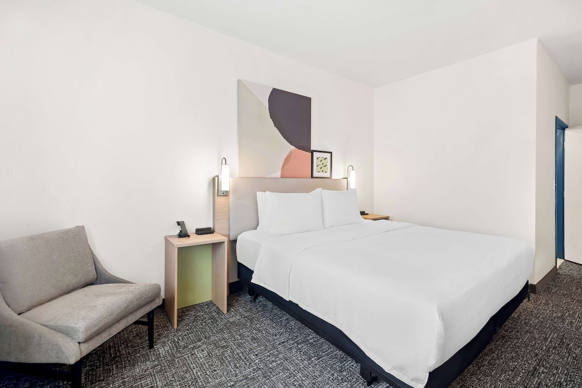 Habitación de hotel con cama, una silla, una mesita de noche y una obra de arte colgada en la pared.