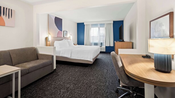 A hotel room with a bed, a desk, a chair, a sofa, a lamp, and a picture frame on the wall. - Spark by Hilton Moline Quad Cities (Moline)