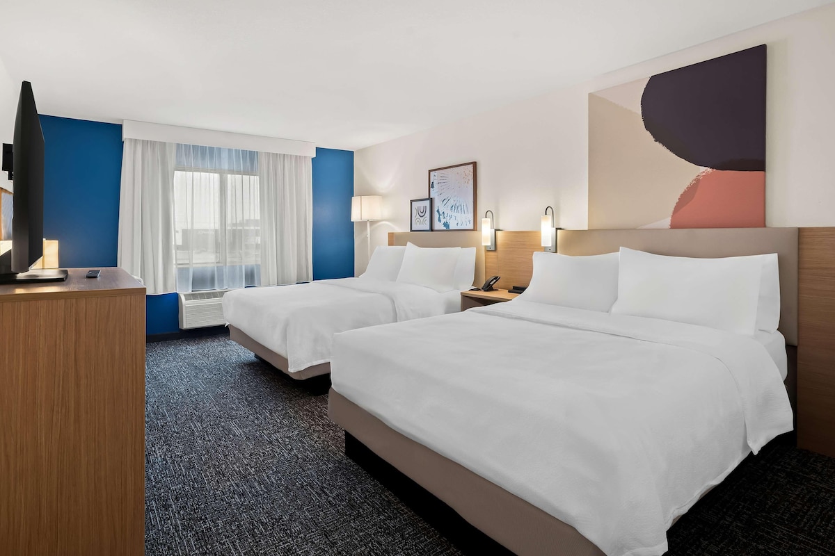 A hotel room with two beds, a desk, a TV, and a large abstract painting on the wall.