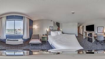 A modern hotel room with a large bed, a desk, a chair, a TV, and a view of the city.