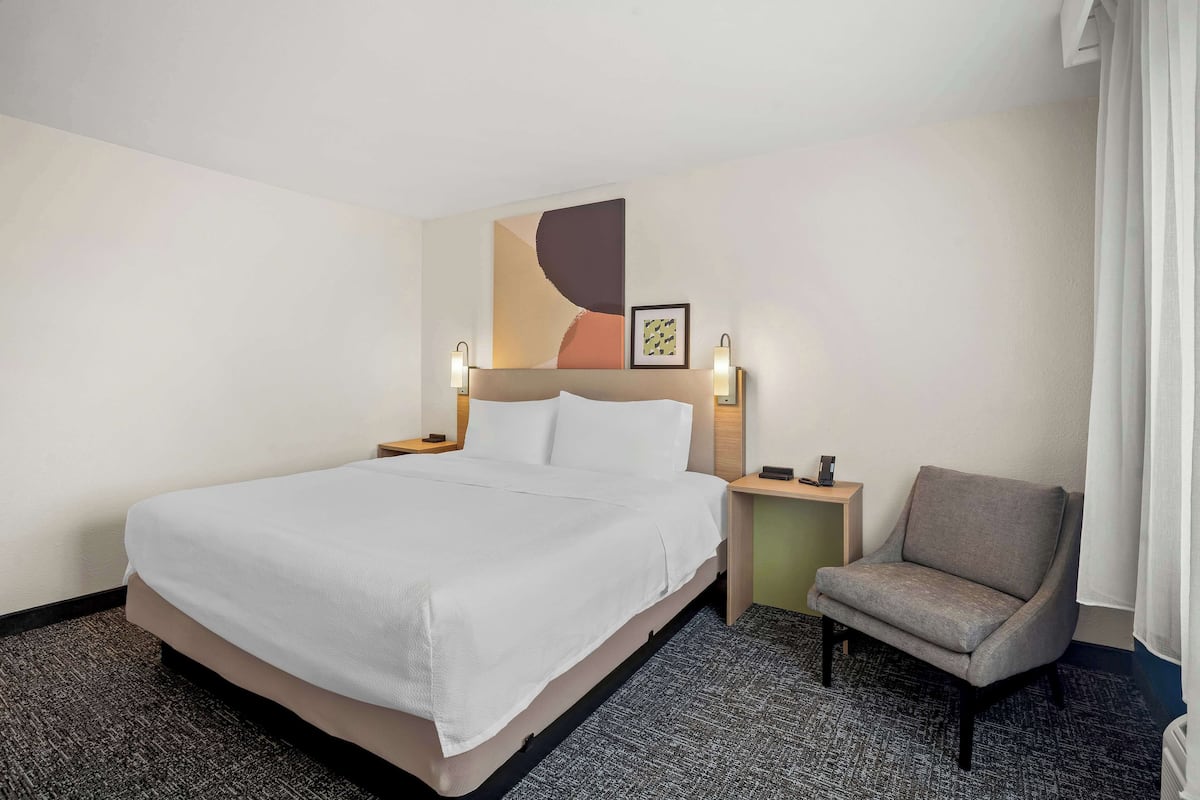 Una habitación de hotel con una cama grande, una silla, una mesita de noche y una obra de arte colgada en la pared.