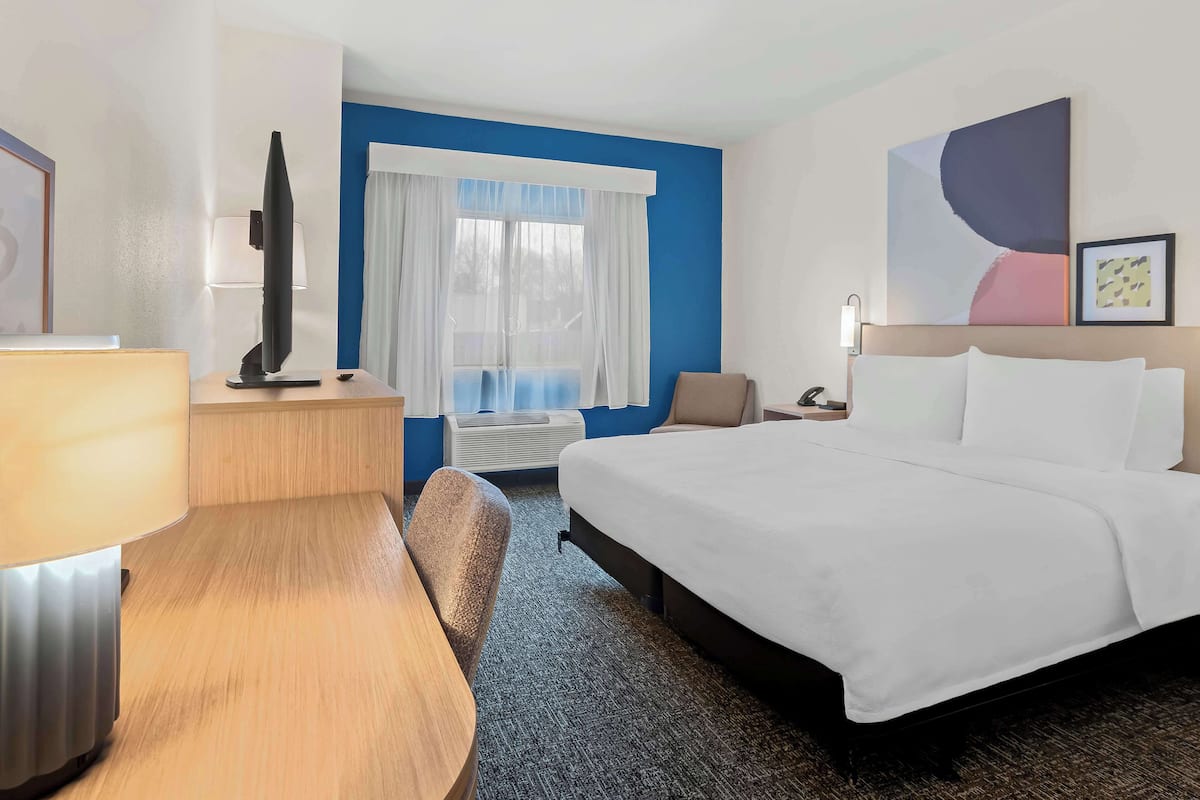 Habitación de hotel con cama, escritorio, silla, televisor, lámpara, ventana con cortinas y cuadros en la pared.