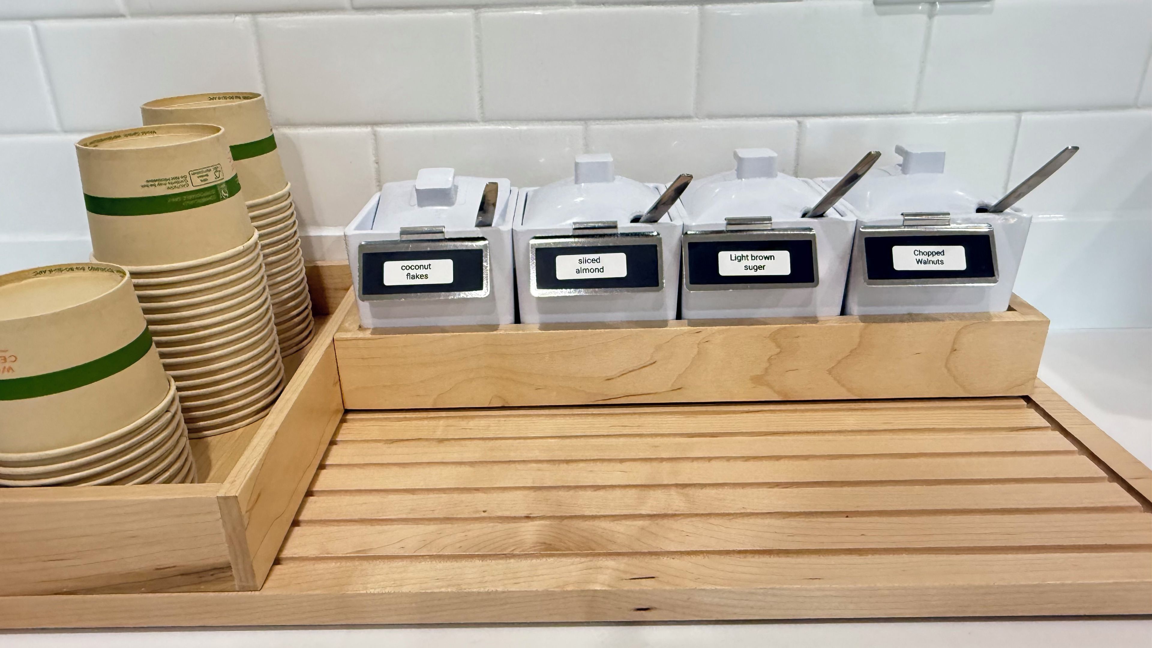 A kitchen counter with labeled containers for coconut flakes, sliced almonds, light brown sugar, and chopped walnuts.