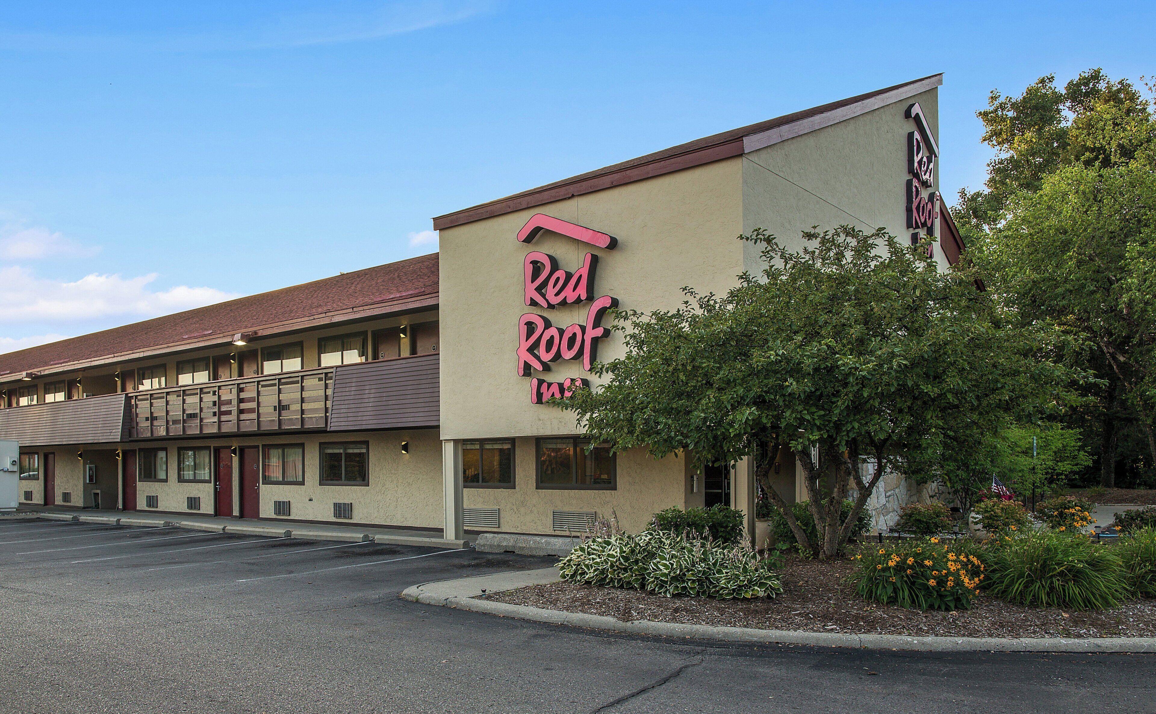 Un hotel Red Roof Inn con un edificio de dos pisos, un estacionamiento y áreas ajardinadas con arbustos y flores.