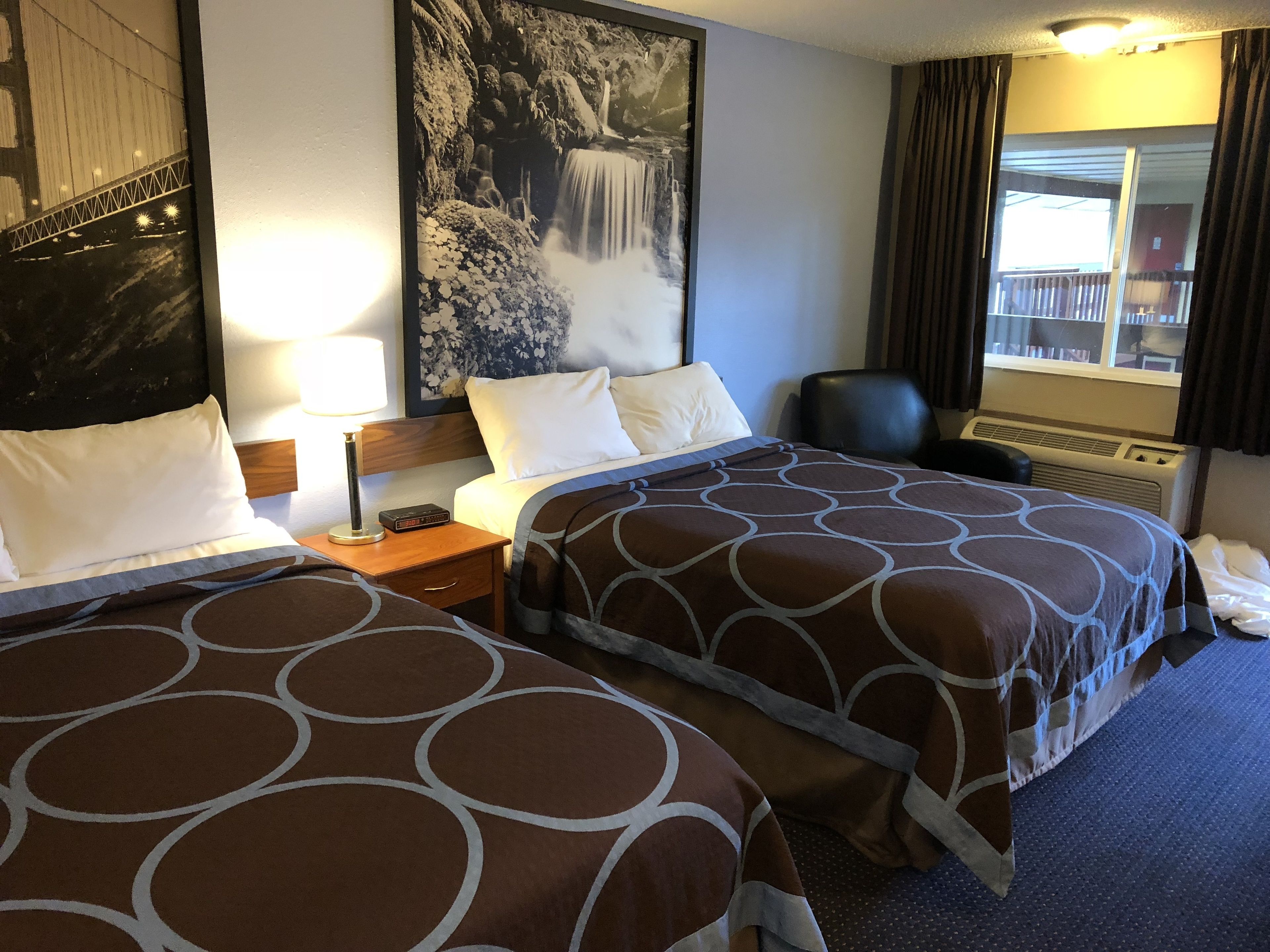 Habitación de hotel con dos camas, una mesita de noche, una lámpara, un cuadro grande en la pared, una ventana con cortinas y vistas a un edificio.
