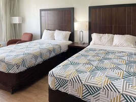 Dos camas dobles con ropa de cama estampada, una mesita de noche con lámpara y una silla en una habitación de hotel.