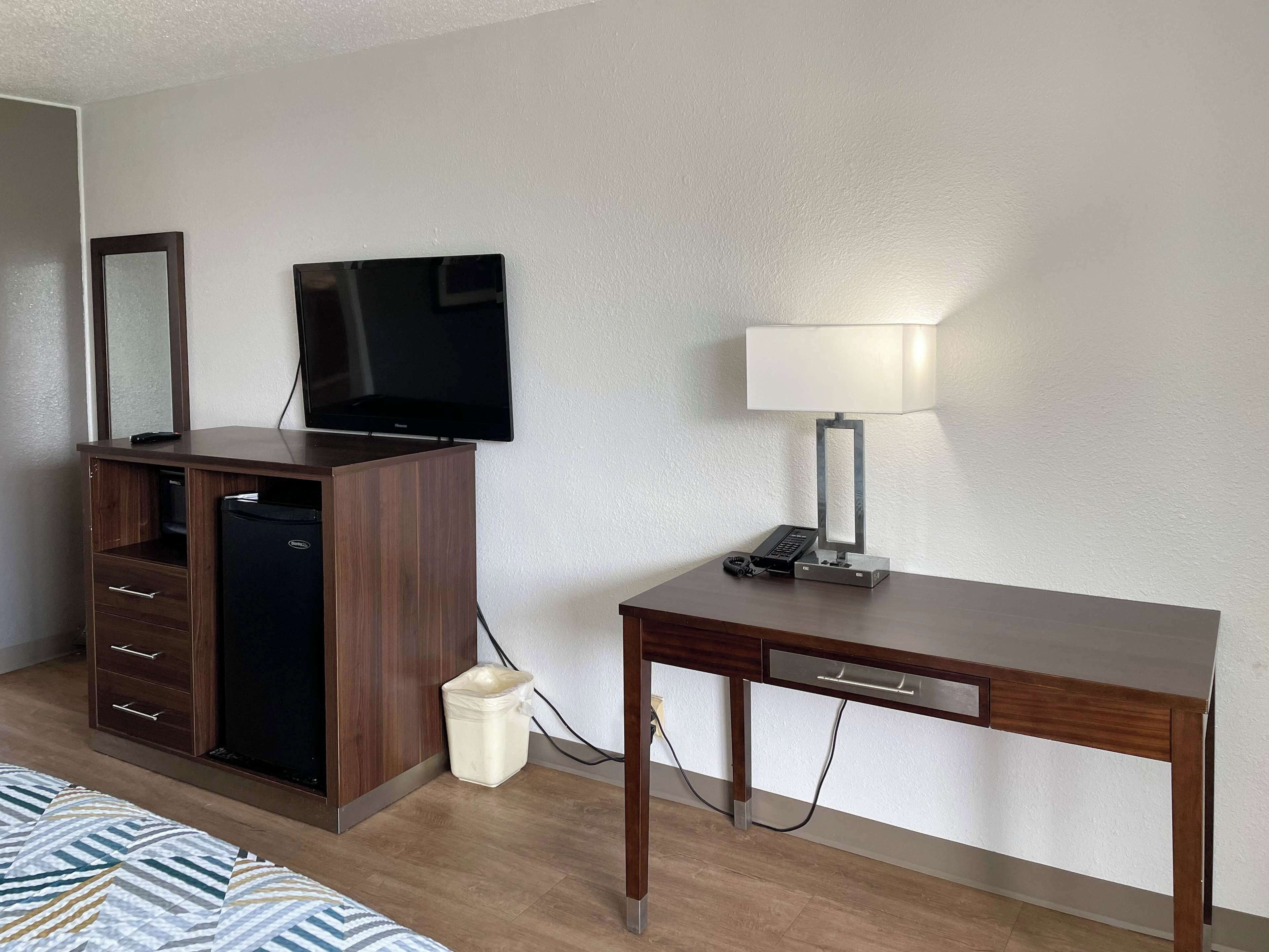 A hotel room with a wooden desk, a television mounted on a cabinet, a trash bin, and a bedside lamp.