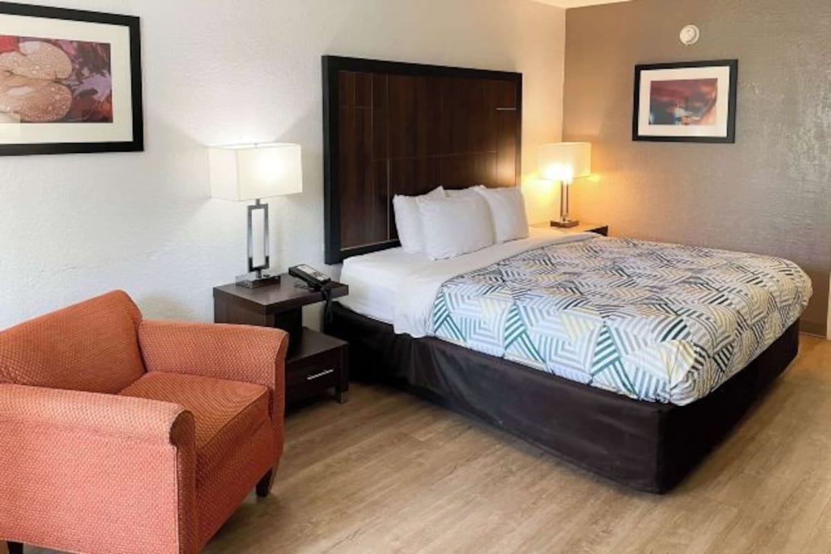 A hotel room with a bed, a nightstand, a lamp, a chair, and two framed pictures on the walls.