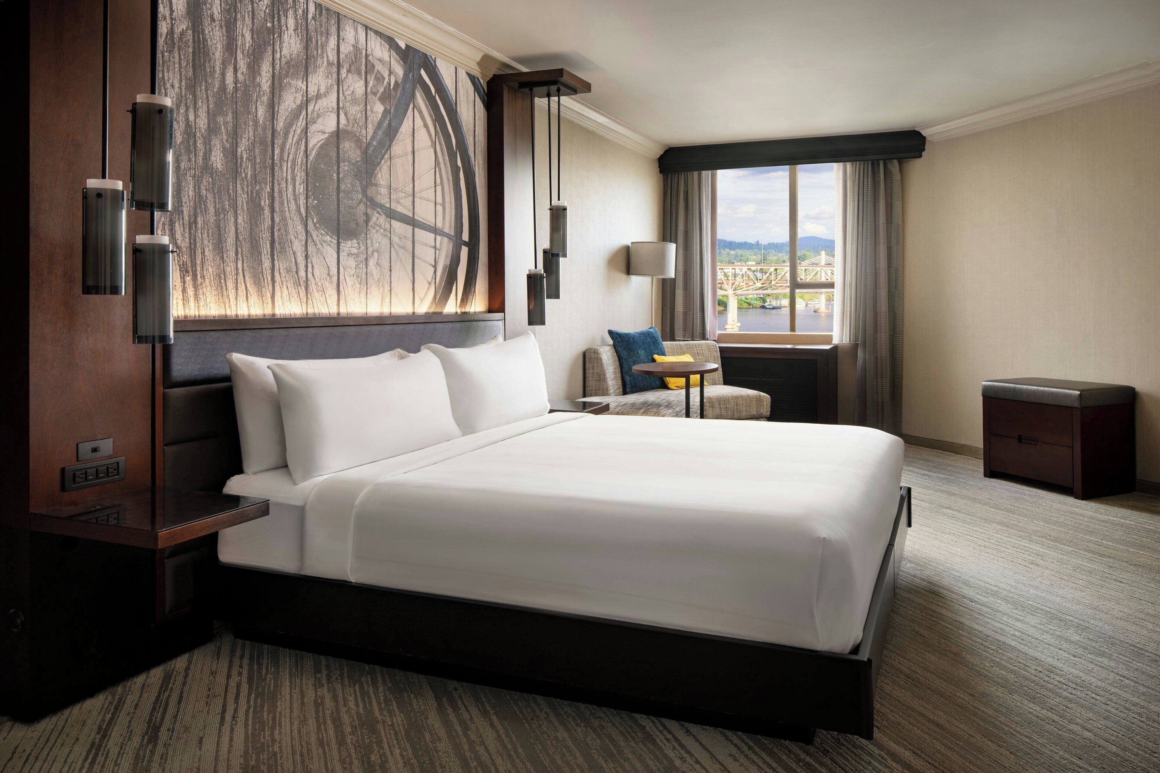 A large bed with white linens, a nightstand, a lamp, a small table with a chair, and a window with a view of a bridge.