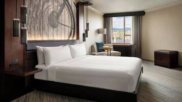 A large bed with white linens, a nightstand, a lamp, a small table with a chair, and a window with a view of a bridge.