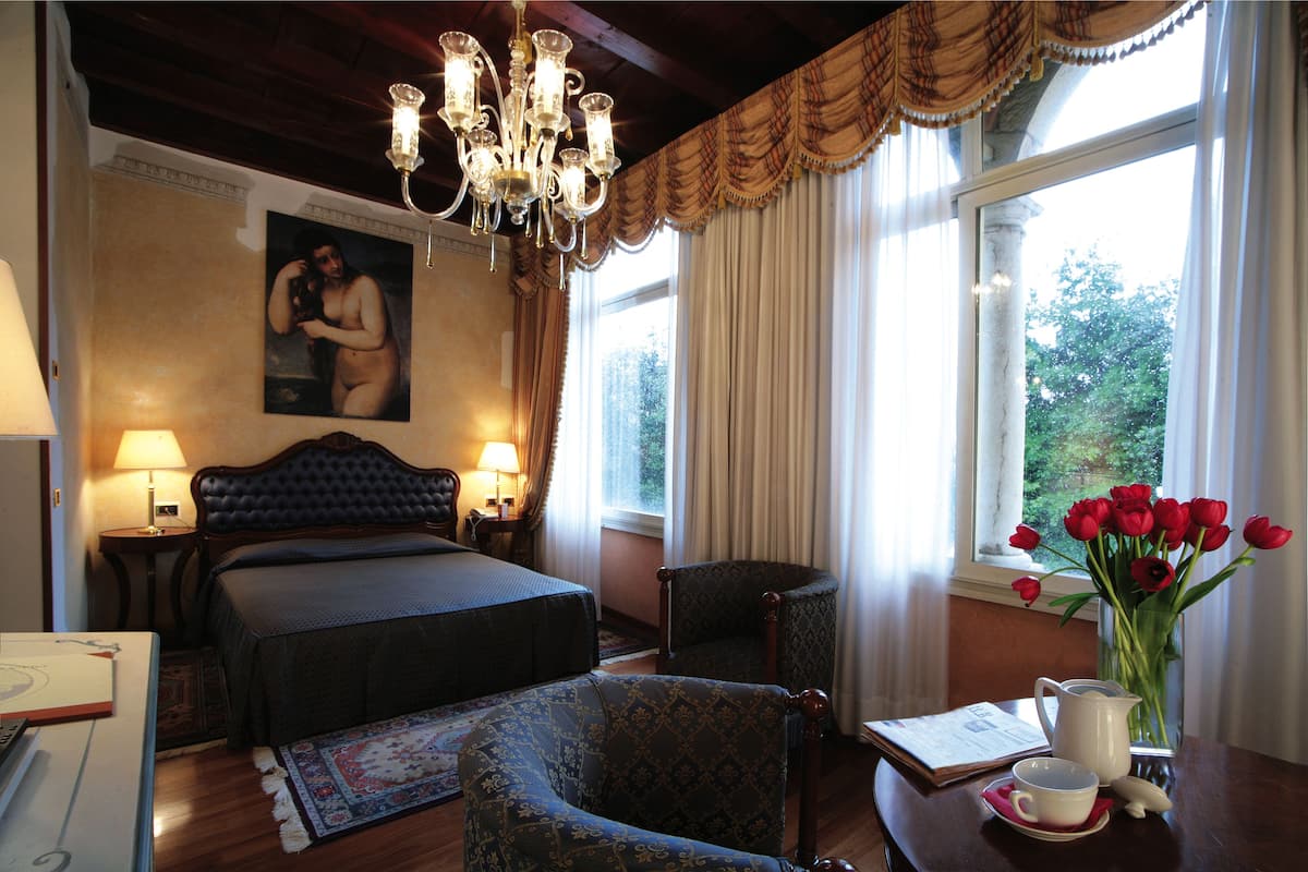 Une chambre d’hôtel avec un grand lit, un coin salon avec un fauteuil, une table agrémentée d’un vase de fleurs et une fenêtre avec des rideaux.