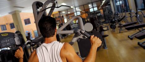 A gym with various exercise machines, a person using a machine, and large windows in the background.