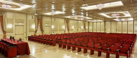 A conference room with rows of red chairs, a long table with microphones, and chandeliers on the ceiling.