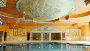 A large indoor swimming pool with a central fountain, surrounded by a classical architectural design with columns and a detailed ceiling.