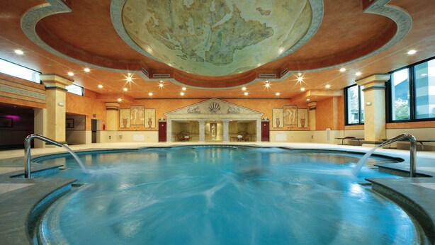 A large indoor swimming pool with a central fountain, surrounded by a classical architectural design with columns and a detailed ceiling mural.