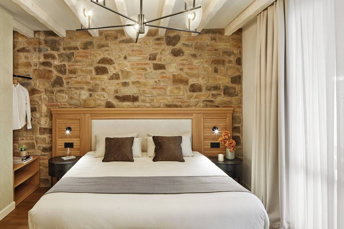 Une chambre avec un mur en pierre, une tête de lit en bois, un grand lit, des tables de chevet et une petite table de nuit ornée d’un vase de fleurs.
