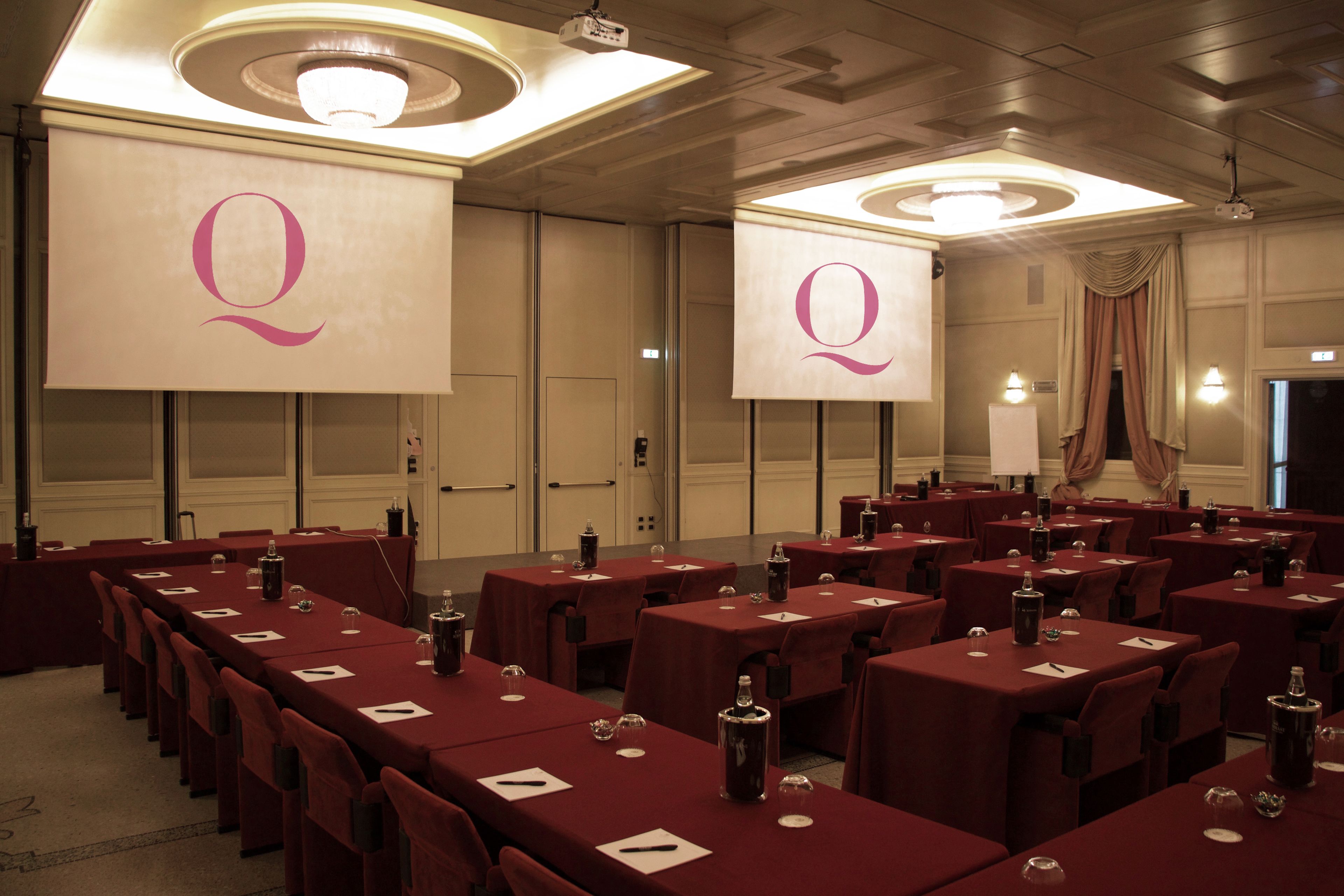 A conference room with multiple tables, each with a red tablecloth and a projector screen displaying a large 'Q'.