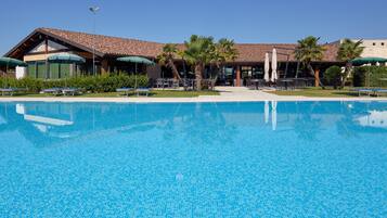 Piscina con sillas de descanso y sombrillas, rodeada de palmeras y un edificio con tejado de azulejos.