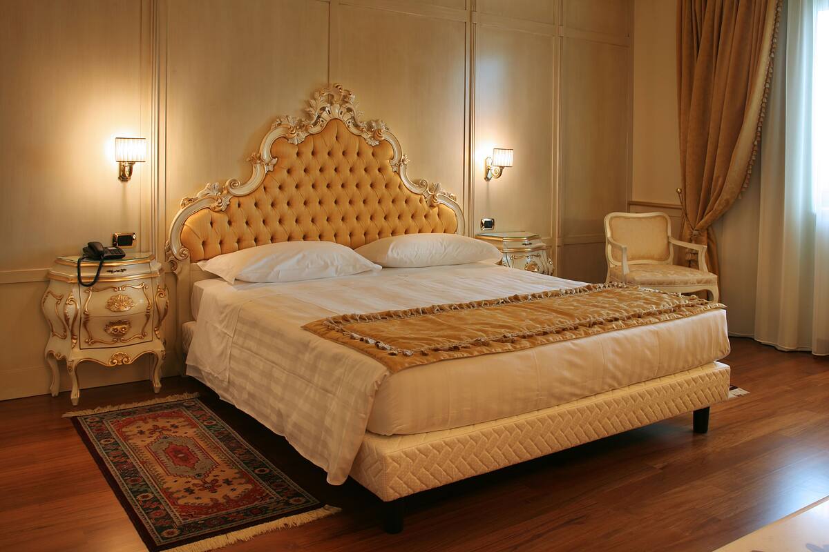 Un grand lit somptueux, doté d’une tête de lit capitonnée, de deux tables de chevet, d’une chaise et d’un tapis à motifs.