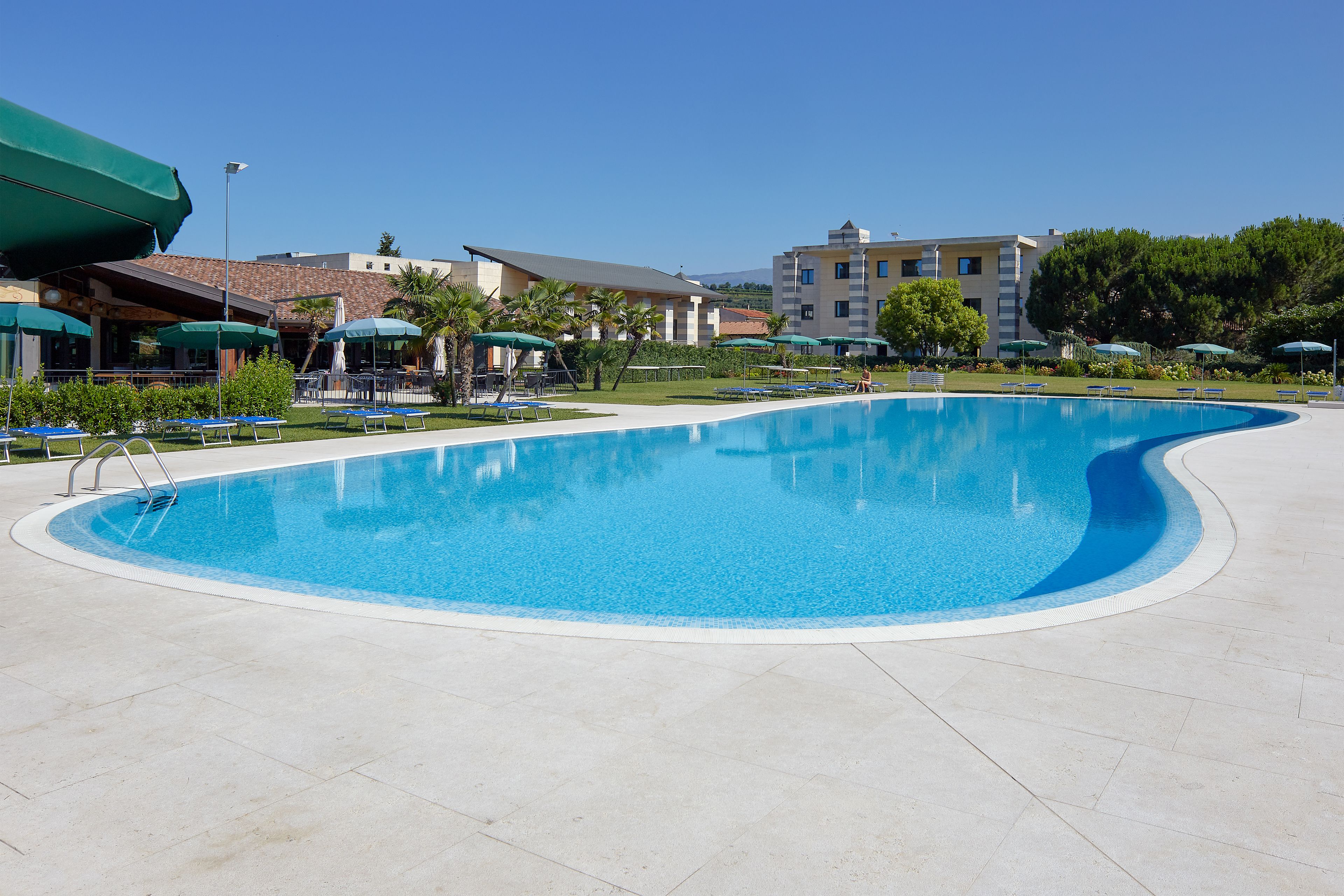 A large, curved swimming pool with a clear blue water surface, surrounded by lounge chairs and umbrellas, with buildings in the background.