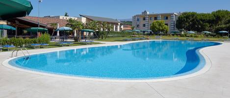 A large, curved swimming pool with a clear blue water surface, surrounded by lounge chairs and umbrellas, with buildings in the background.