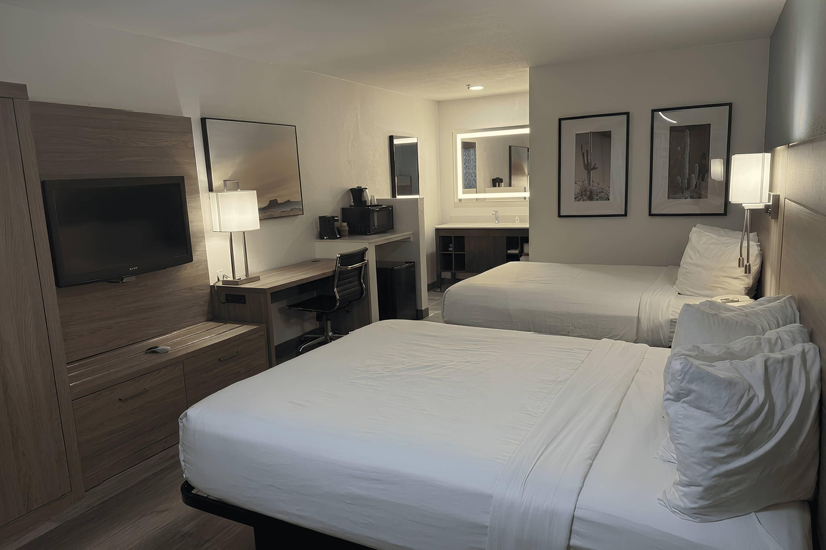 Une chambre d’hôtel avec deux lits, un bureau avec un ordinateur, une télévision et une salle de bain équipée d’un miroir et d’un lavabo.