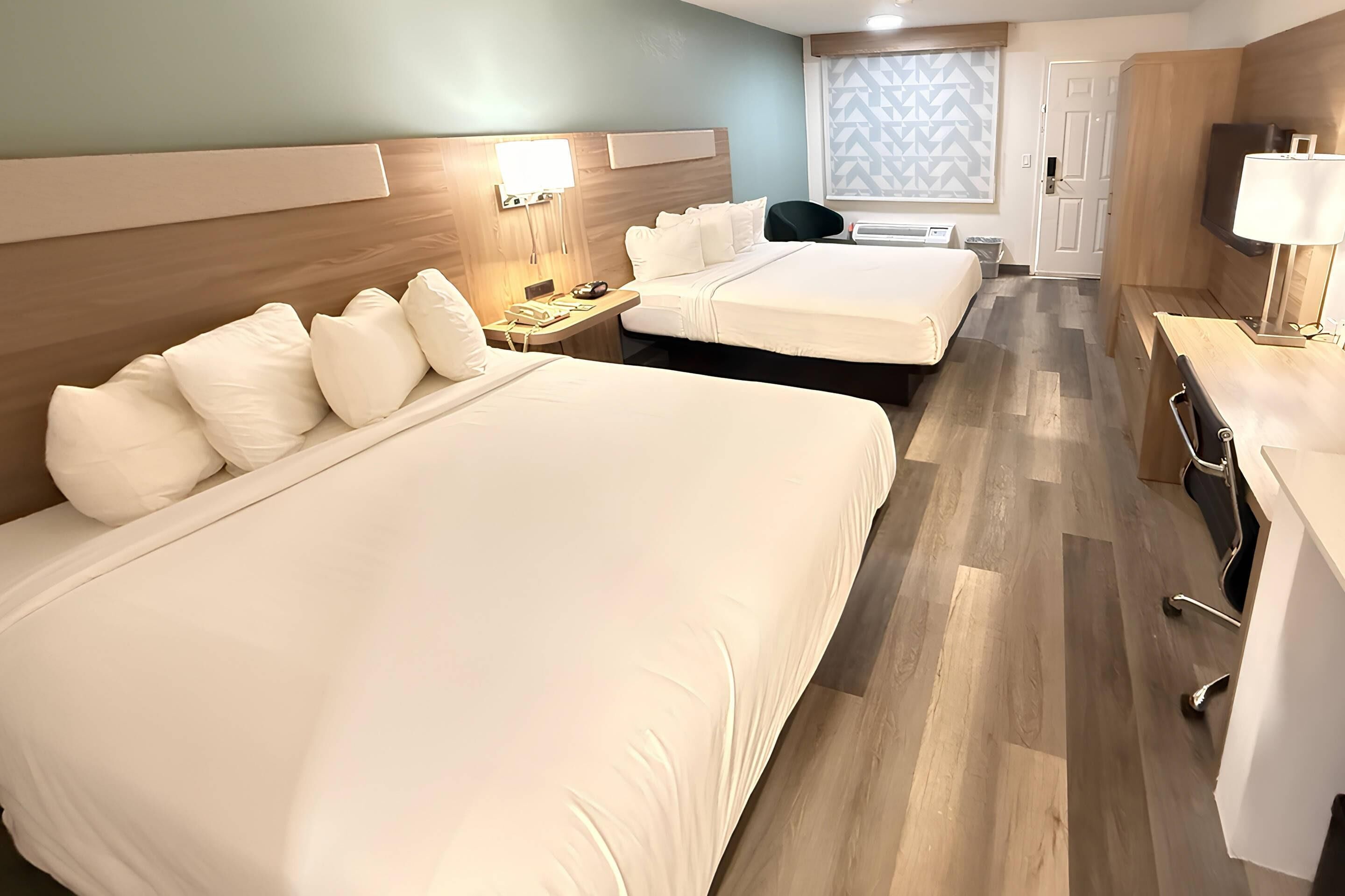 Habitación de hotel con dos camas, un escritorio, una silla, una mesita de noche y una ventana con diseño geométrico.