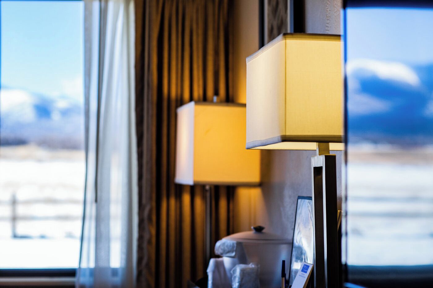 A hotel room with a large window offering a view of a snowy landscape. There are two lamps on the bedside table.