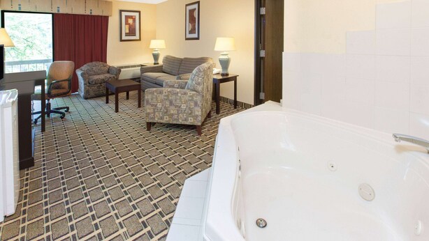 A hotel room with a large bathtub, two armchairs, a desk with a computer, and a window with a red curtain. - Days Inn & Suites by Wyndham Madison Heights MI (Madison Heights)