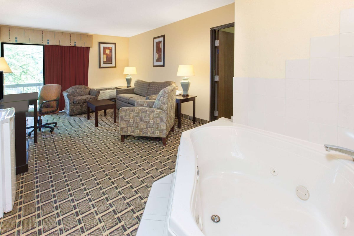 A hotel room with a large bathtub, two armchairs, a desk with a computer, and a window with a red curtain.