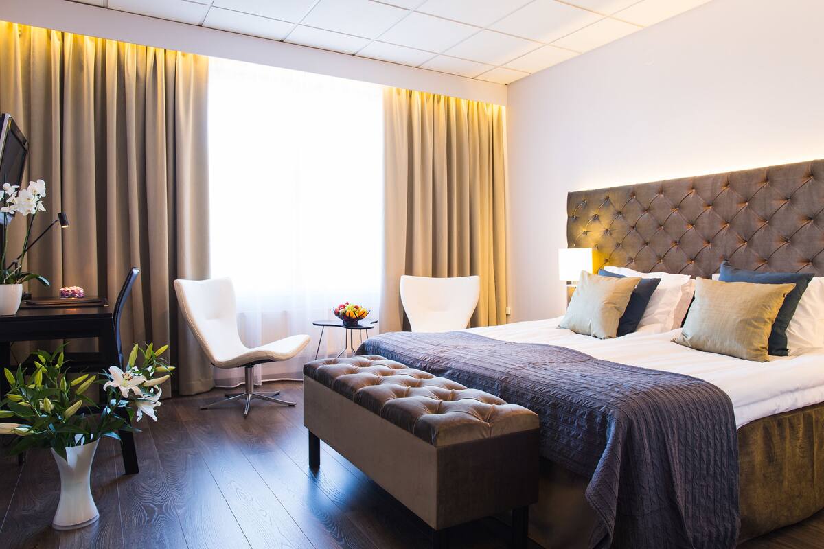 A hotel room with a large bed, a sitting area with a chair, a small table with a bowl of fruit, and a potted plant.